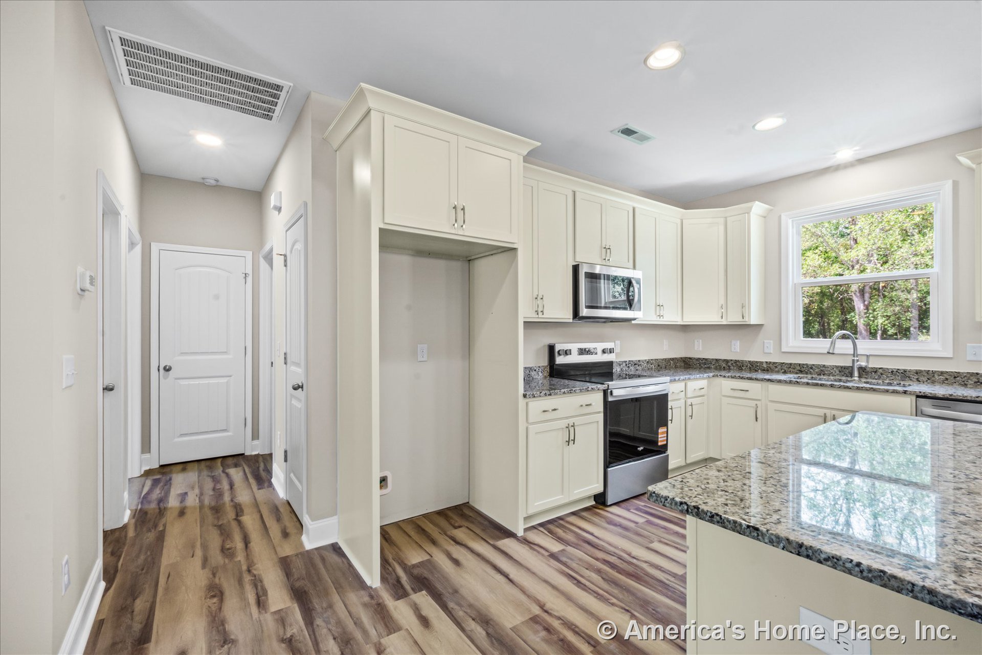 Cream kitchen cabinetry with granite countertops, stainless steel built-in microwave and electric oven, wood-look vinyl plank flooring, recessed ceiling lights, large window above