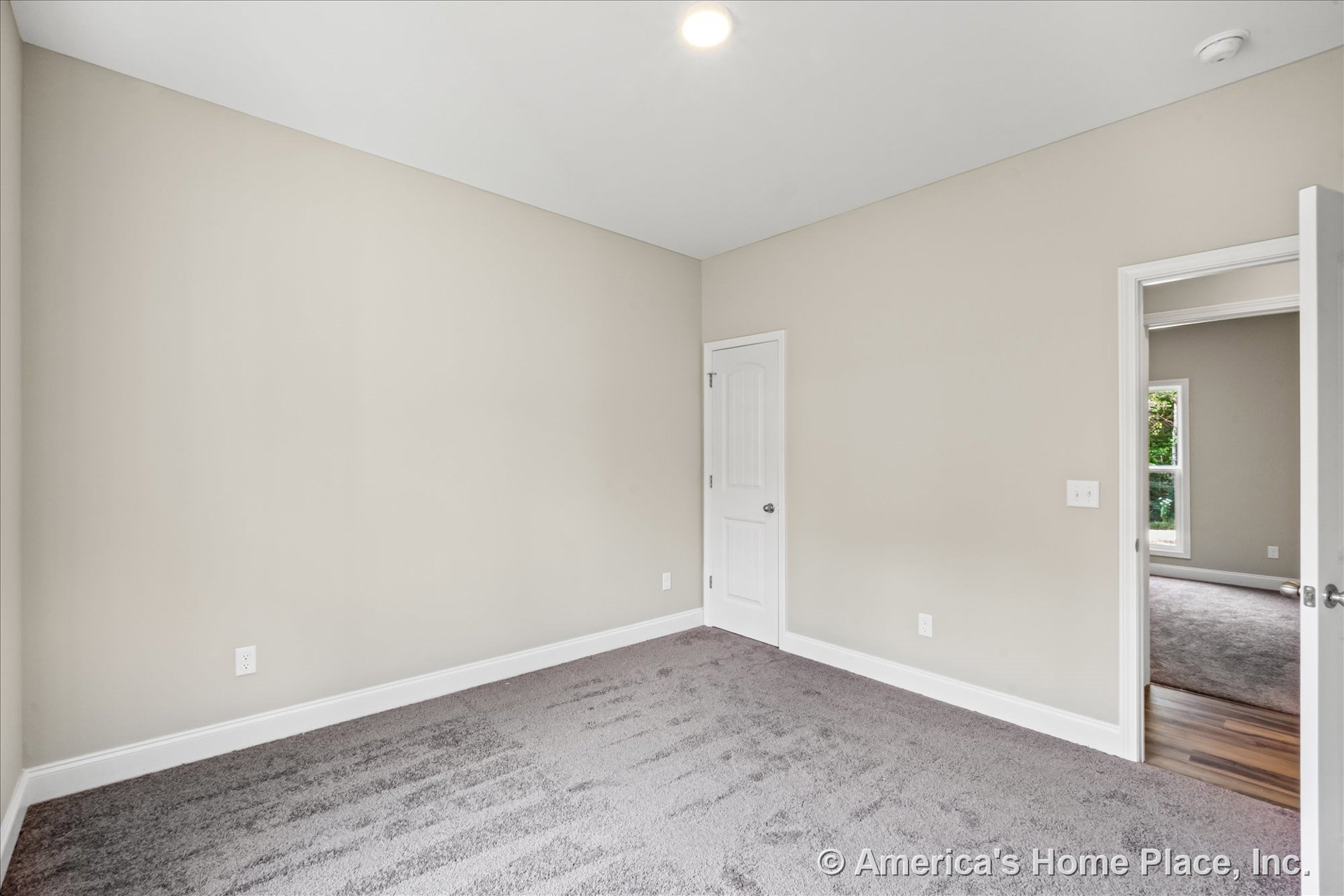 Bedroom with light beige walls, white baseboards and trim, gray carpet flooring, flush ceiling light, white paneled door, and view into adjacent room with window.