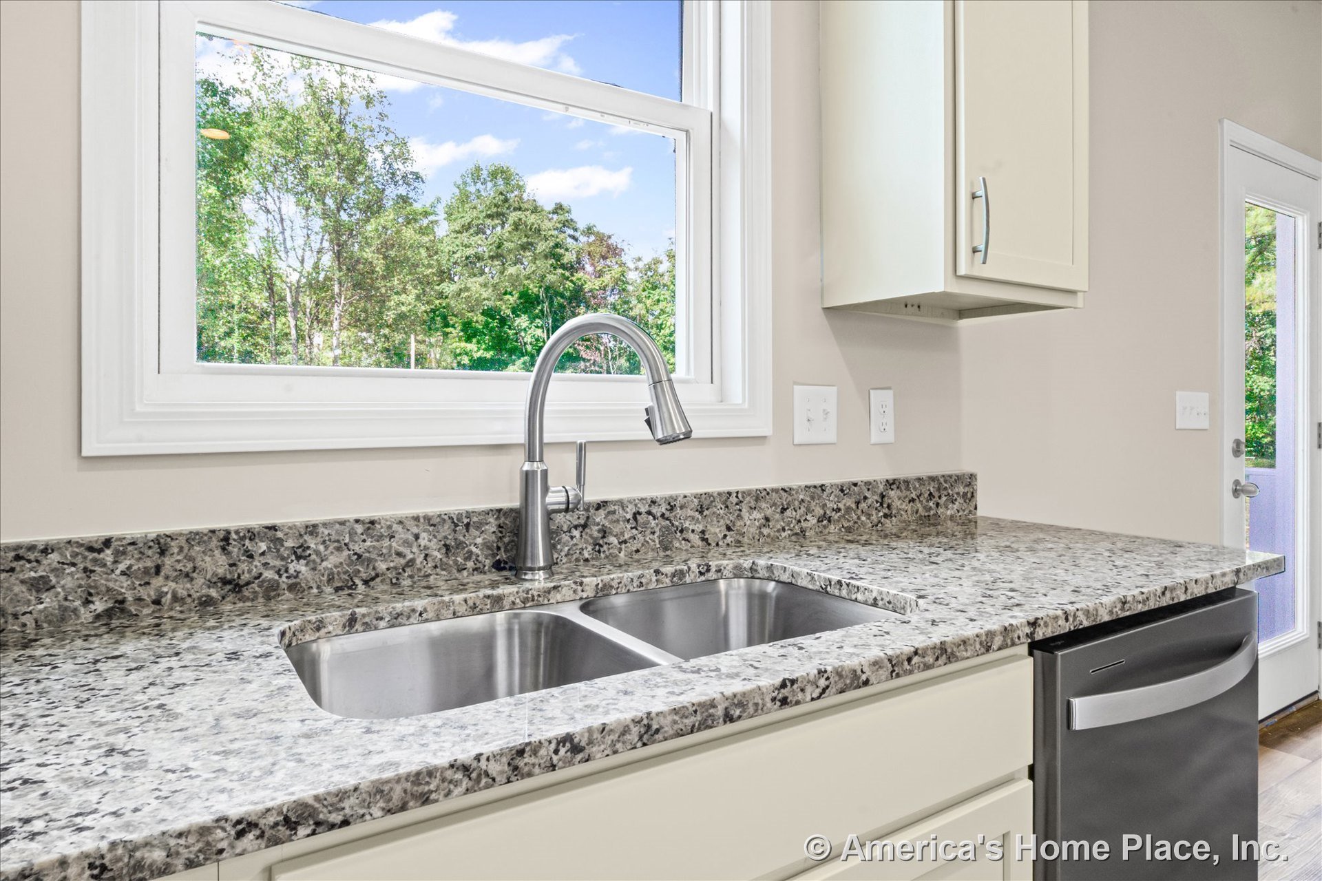 Granite countertop with double stainless steel sink and gooseneck faucet beneath a large window with white trim, cream cabinetry, and built-in dishwasher.