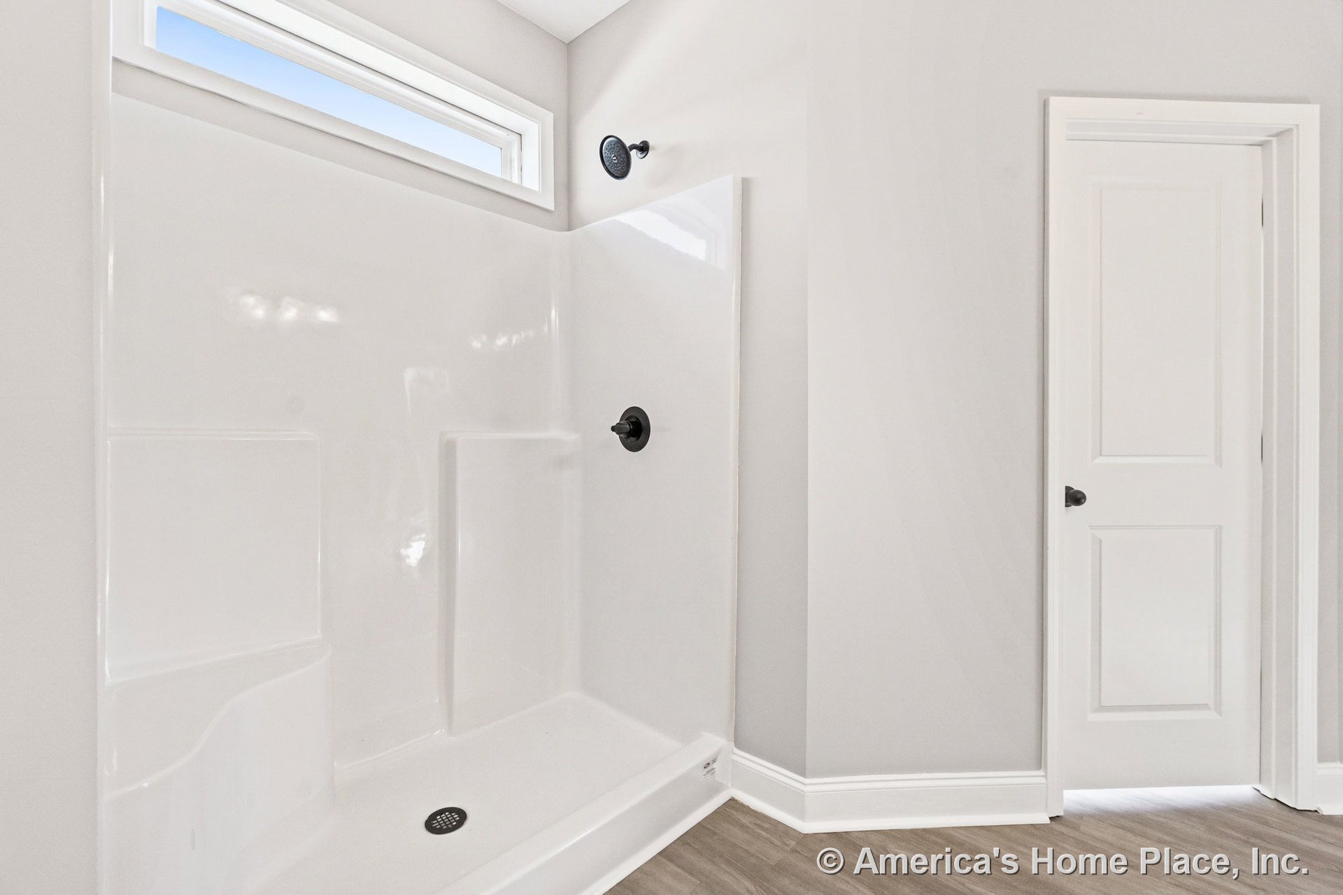 Walk-in shower with built-in shelves, matte black fixtures, and a horizontal transom window; light wood-look flooring, white paneled door, and white baseboard trim.