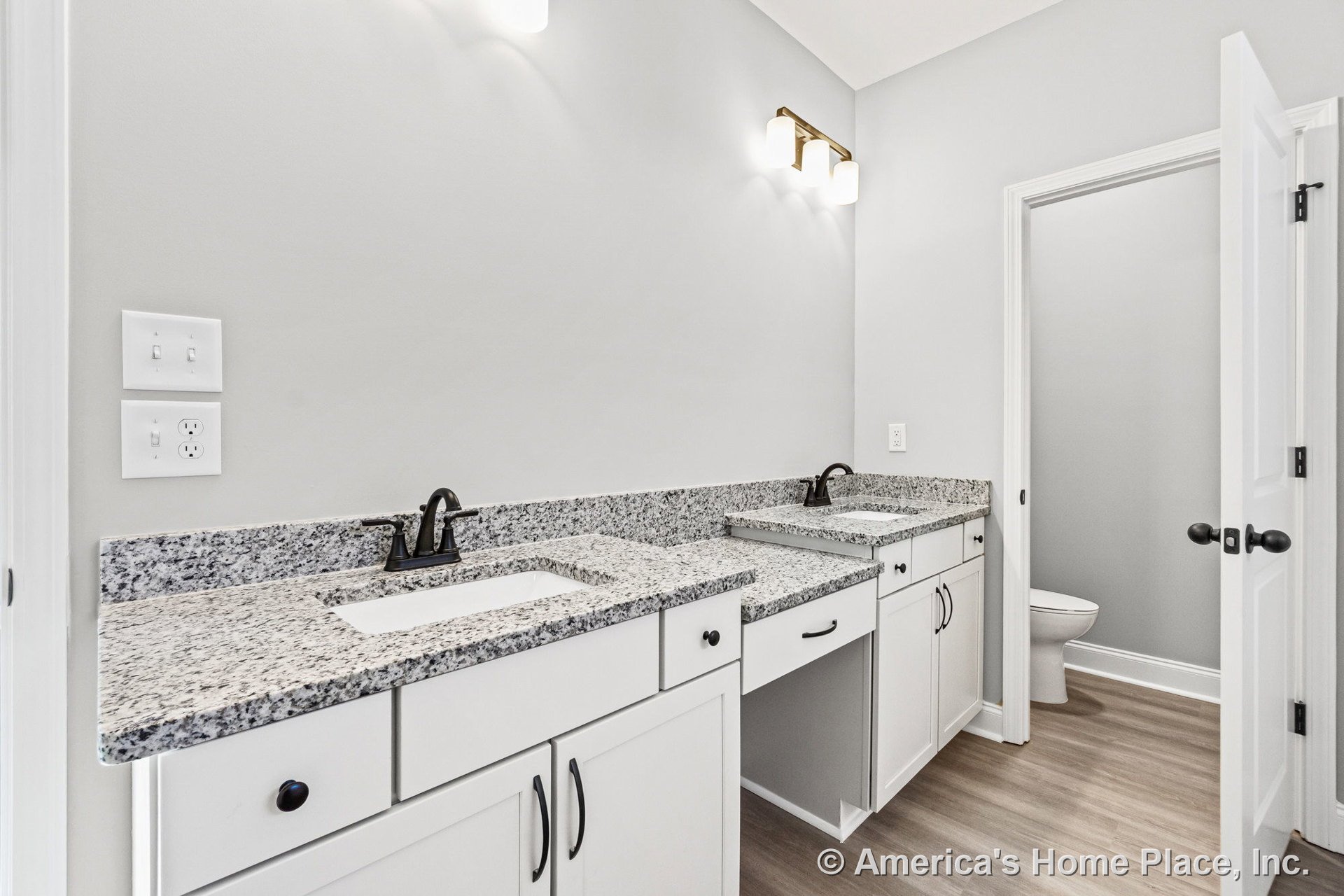 Double vanity with granite countertops, white shaker cabinets, matte black faucets, wall-mounted light fixtures, light wood-look flooring, and a separate toilet area.
