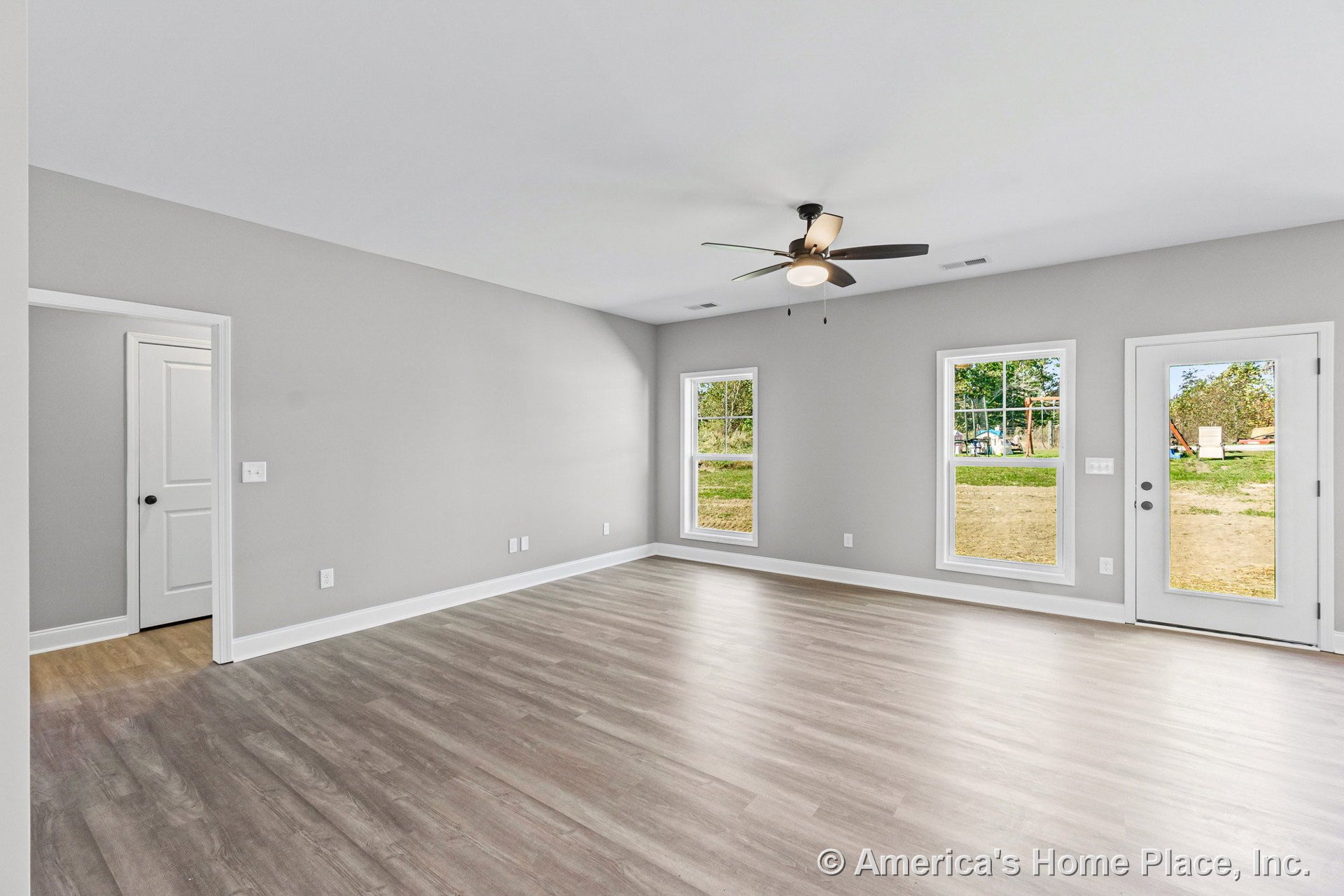 Open living area with light wood-look vinyl plank flooring, gray painted walls, white trim and baseboards, ceiling fan with light fixture, three large windows, and glass exterior