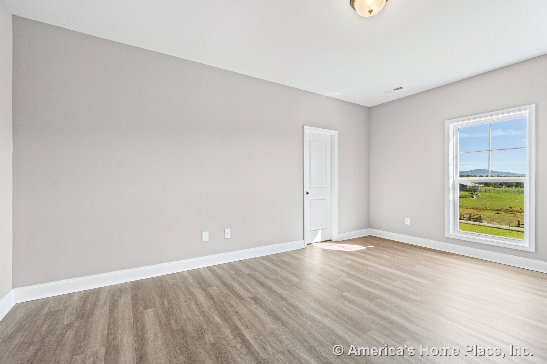 Bright bedroom with light wood-look flooring, large window framed by white trim, neutral painted walls, white baseboards, paneled door, and flush mount ceiling light.