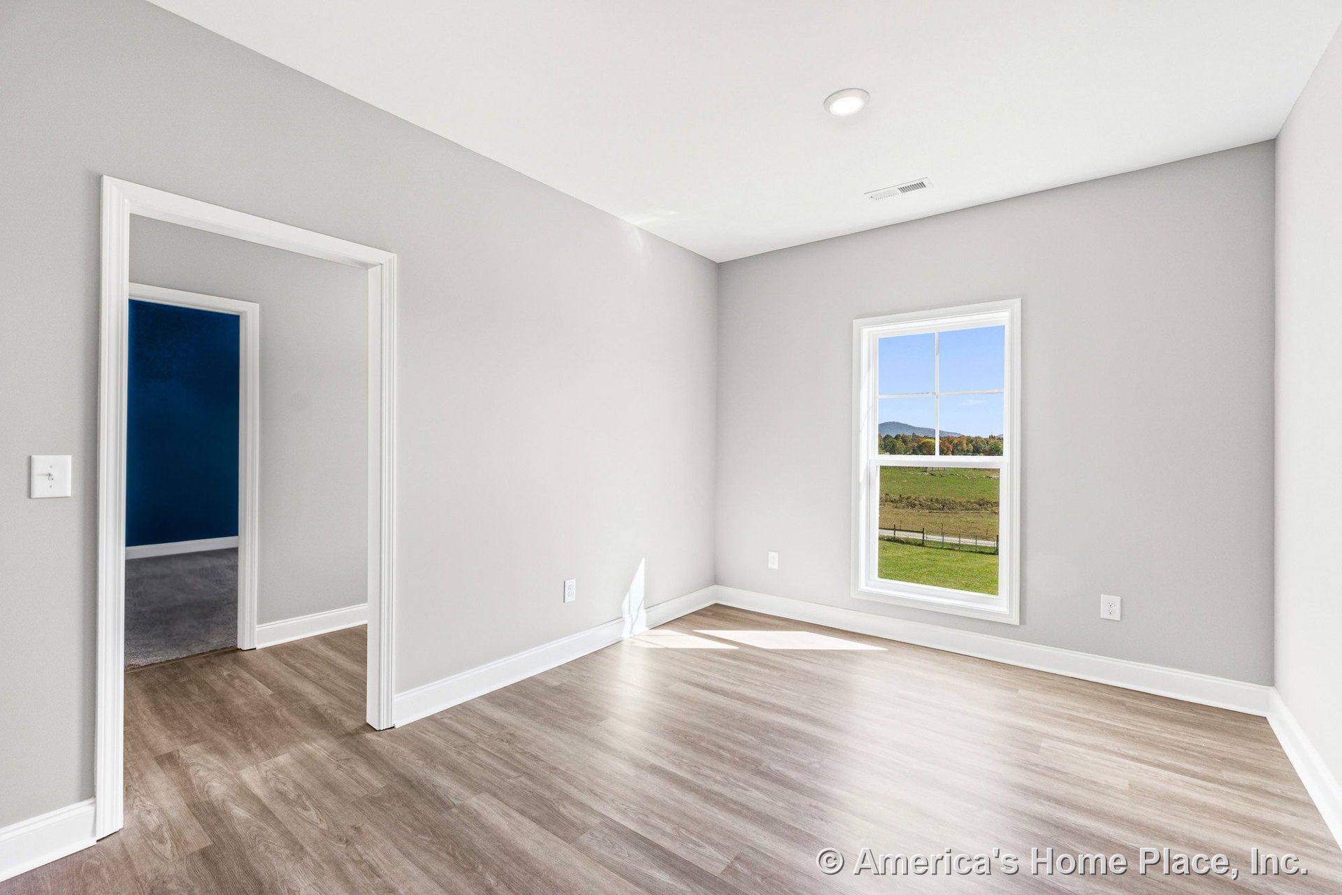 Bright bedroom with light wood-look vinyl plank flooring, large window framed in white trim, smooth gray walls, white baseboards, recessed ceiling light, and open doorway leading