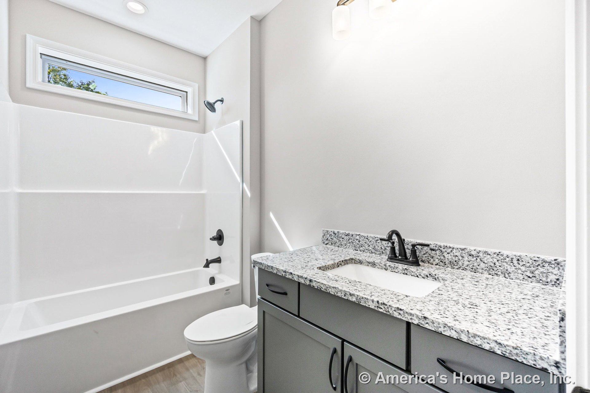 Built-in white acrylic bathtub beneath a horizontal transom window, granite vanity countertop with matte black faucet fixtures, gray shaker-style cabinetry, wall-mounted vanity