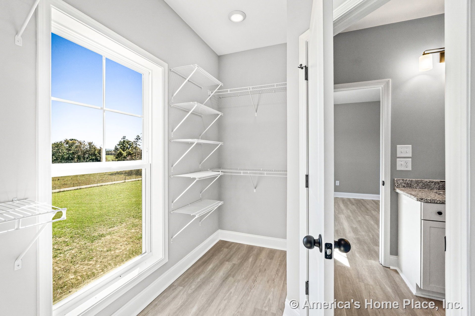 Walk-in pantry with wire shelving, large window framed in white trim, light wood flooring, gray painted walls, recessed ceiling light, white cabinetry topped with granite