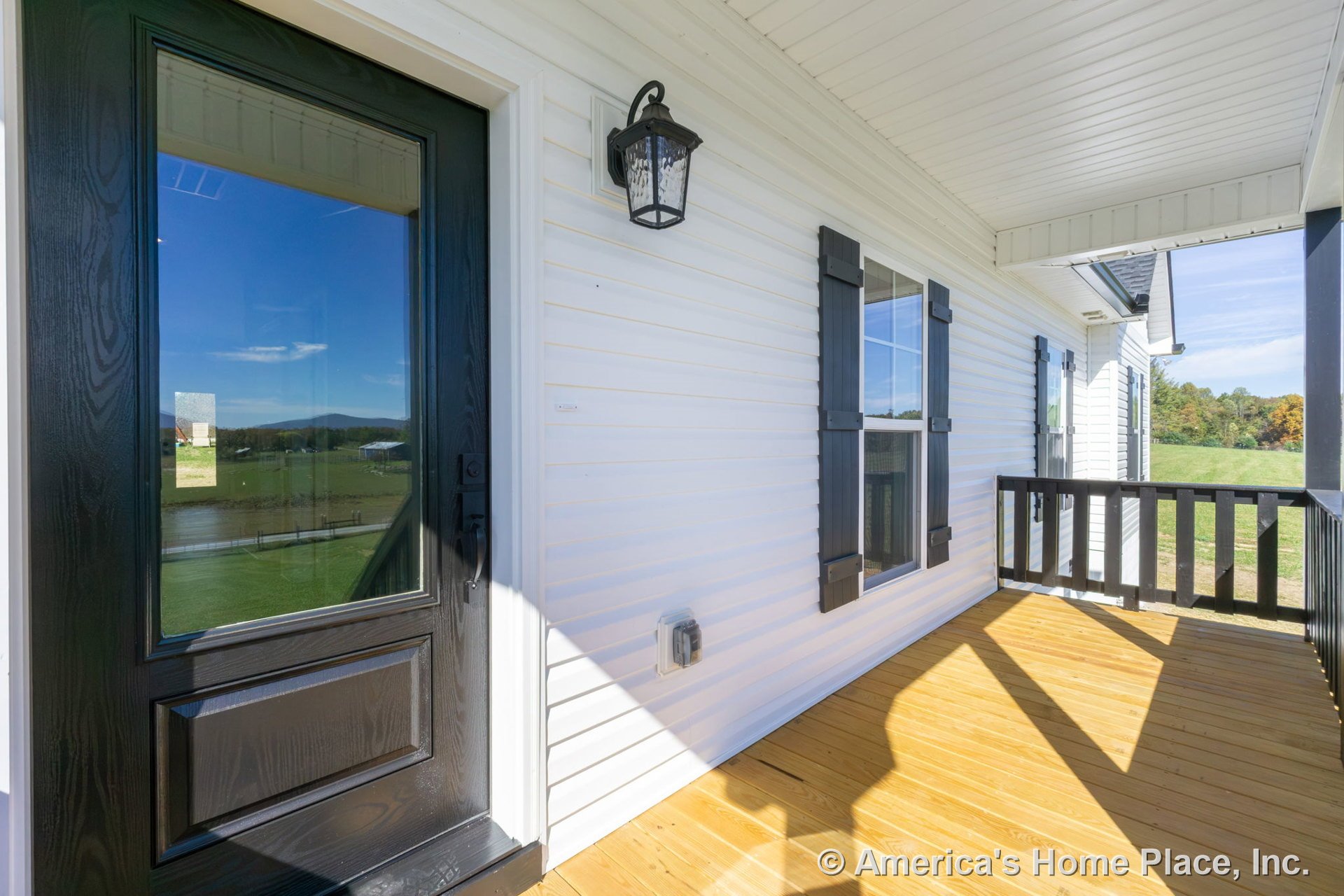 Black framed glass door with sidelights set in white vinyl siding, black window shutters, covered porch ceiling, wood plank flooring, black exterior lantern, double hung windows