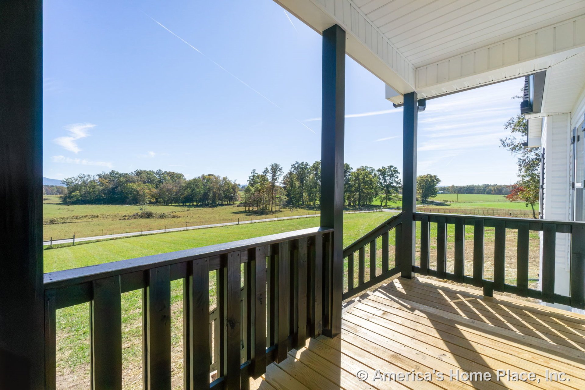 Covered porch with wood plank flooring, black vertical railings, white ceiling, structural support columns, and exterior siding overlooking open rural fields.