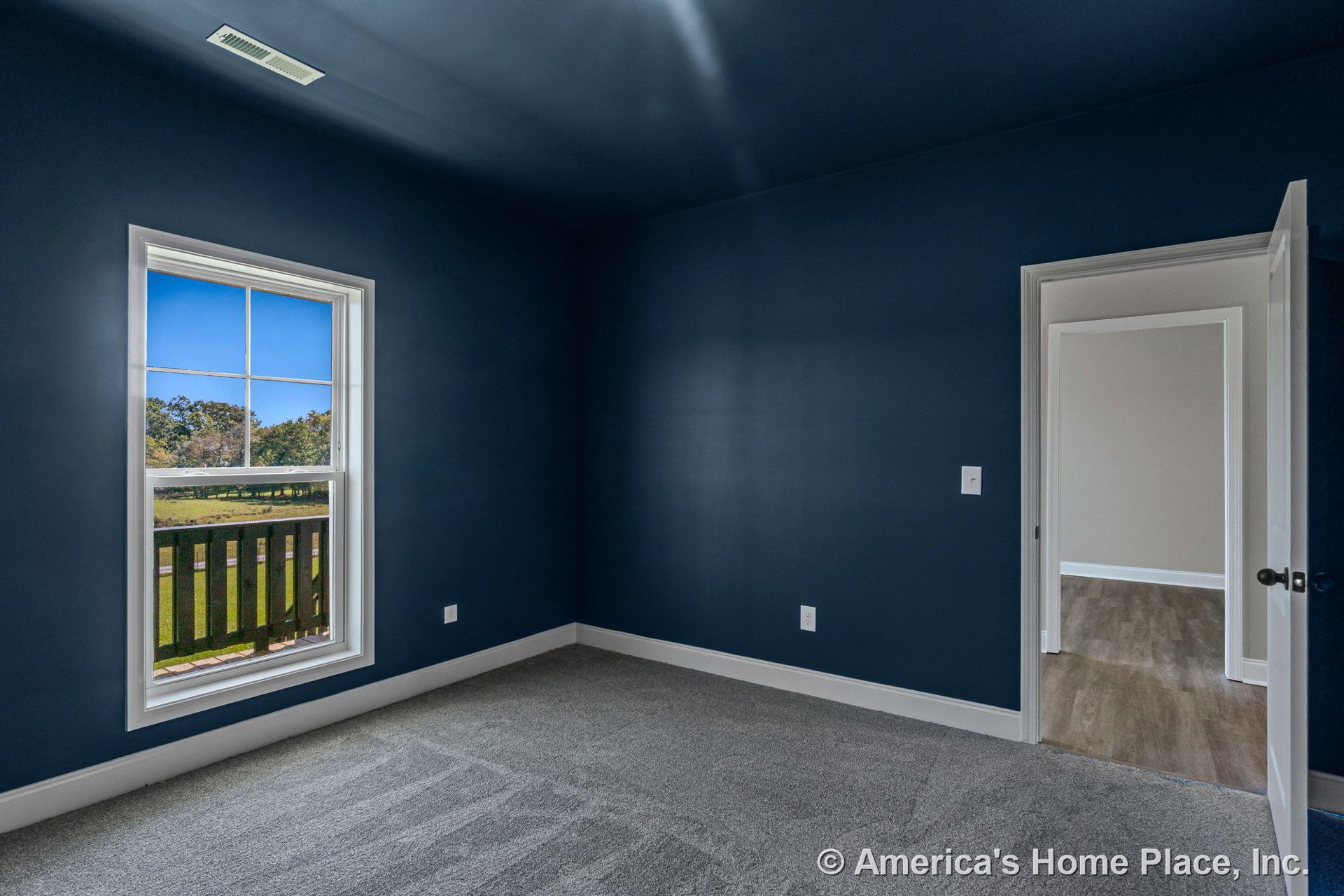 Deep blue walls with white baseboards and trim, large window with muntins allowing natural light, gray carpet flooring, open doorway to adjacent room, and ceiling vent in a modern