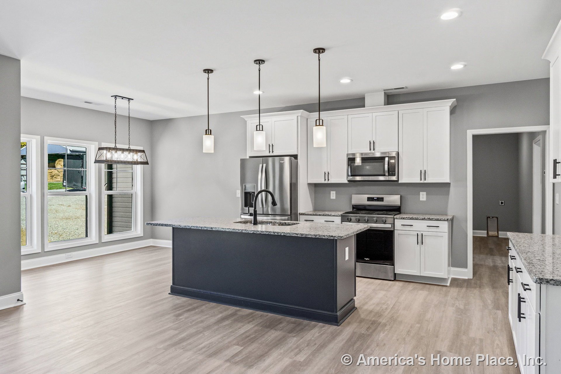 Spacious kitchen with large central island featuring sink, three pendant lights overhead, white shaker cabinets, granite countertops, stainless steel refrigerator, oven, built-in