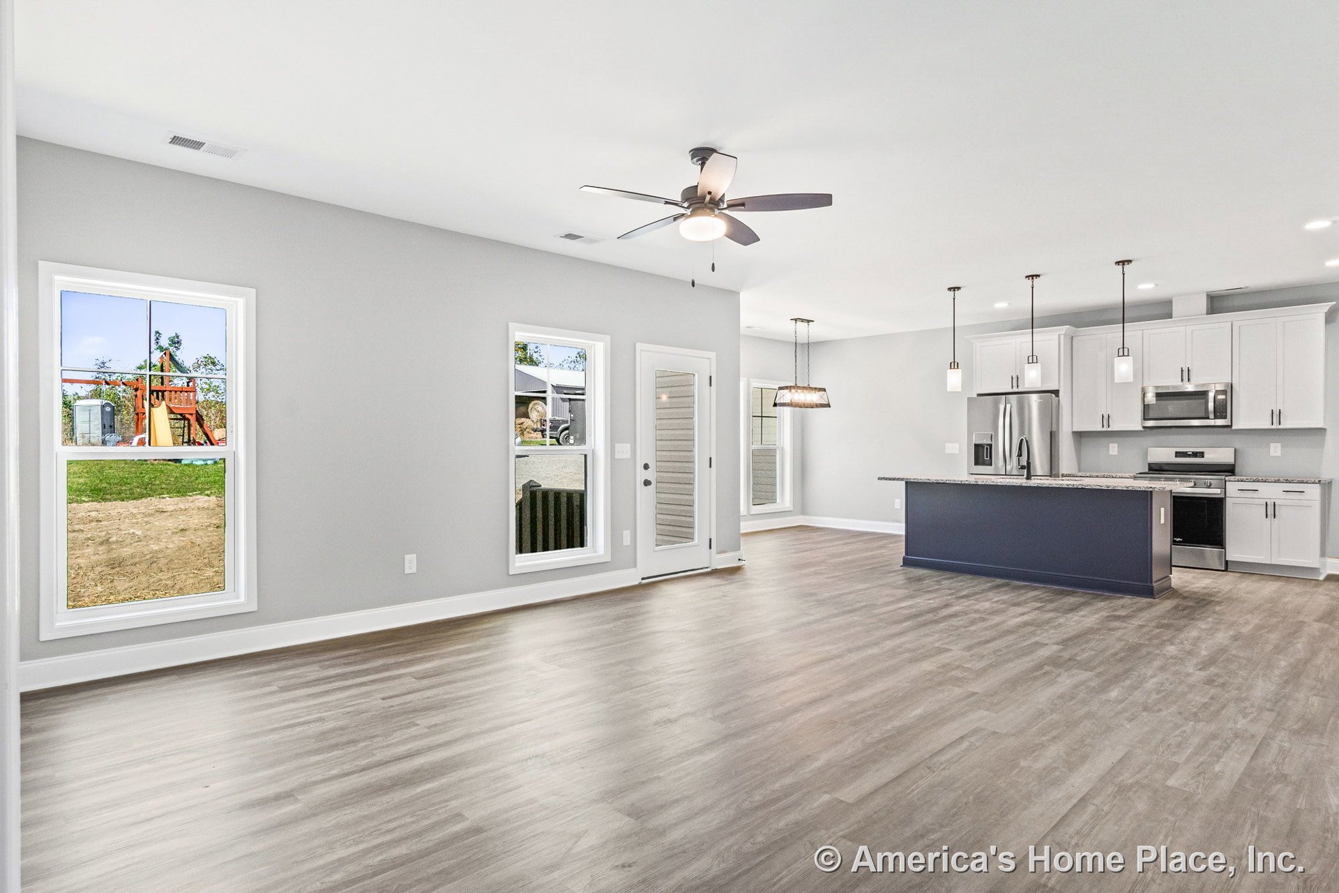 Open-concept living area with large windows, gray wood-look flooring, ceiling fan with light, modern kitchen featuring white cabinets, granite countertops, blue kitchen island