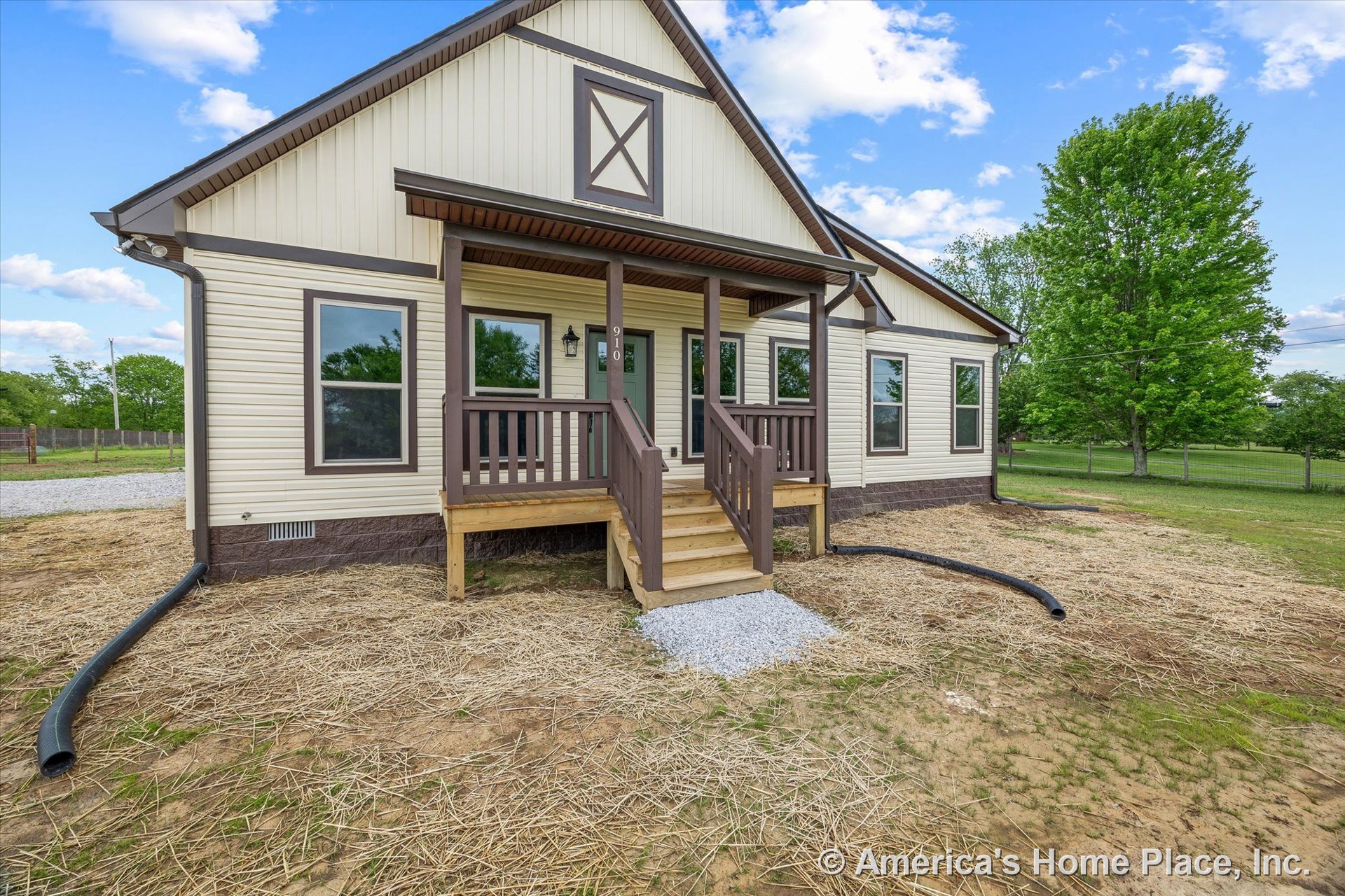 Covered front porch with wood railings and steps, vinyl siding exterior, multiple rectangular windows with brown trim accents, gravel entry pad, front door, and exterior lighting