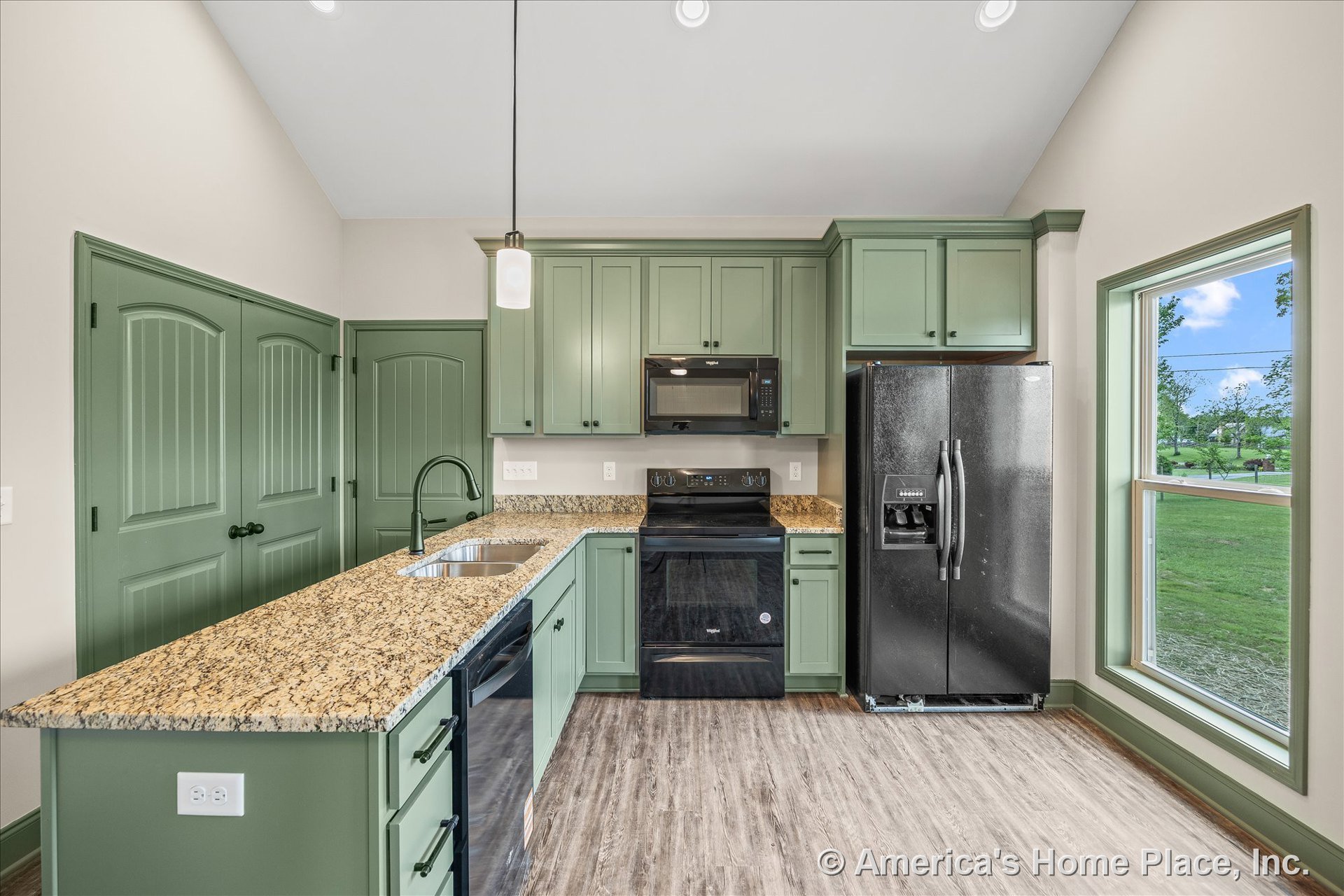 Green shaker cabinets with granite countertop peninsula, black refrigerator and range, double basin undermount sink beneath large window with green trim, pendant ceiling light
