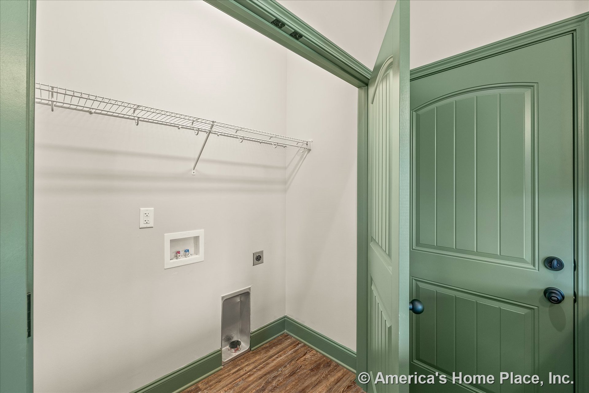 Laundry room with green painted trim and door, wire shelving above washer and dryer hookups, wood-look flooring, utility outlet and plumbing box, baseboards, and door hardware.