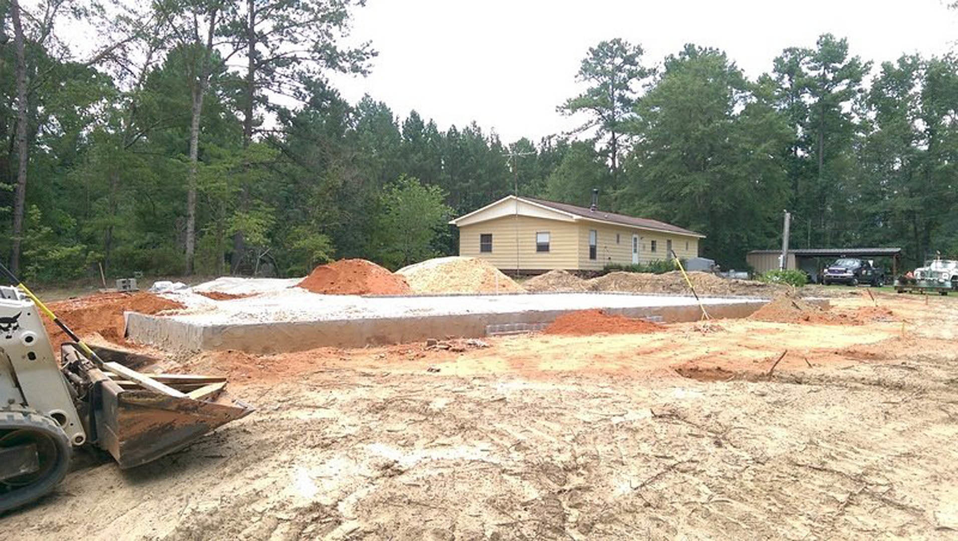 Two-story house with light siding and brick chimney, surrounded by dirt piles and trees, bulldozer parked nearby, car partially visible, roadside in foreground