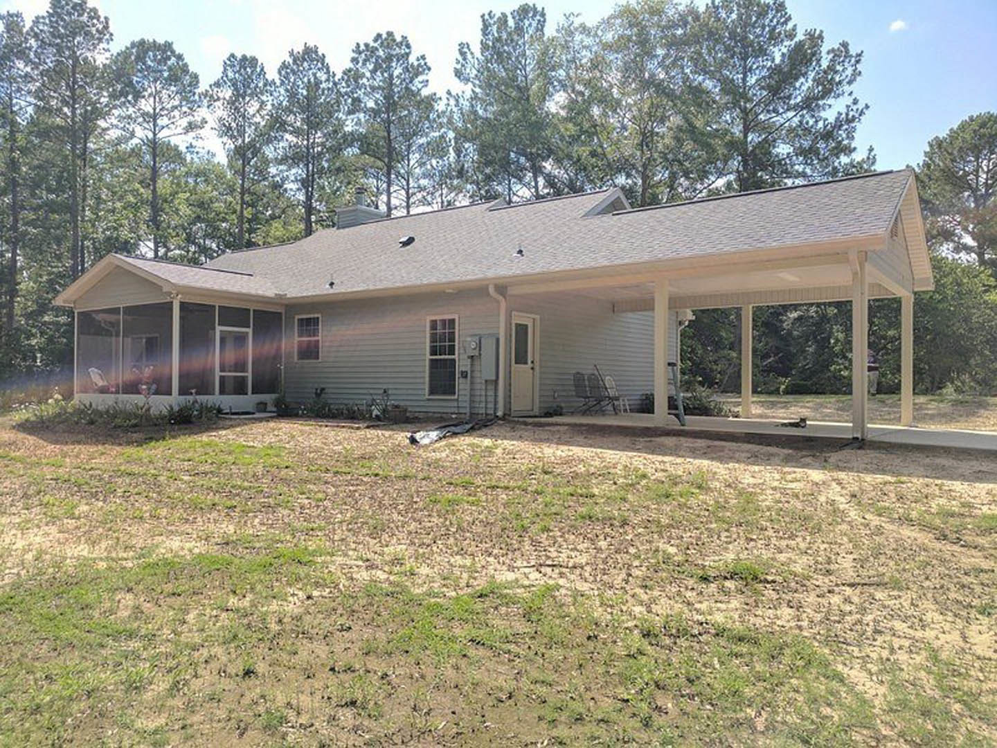 White house with multi-pane windows, covered front porch supported by columns, dirt yard with patches of grass, leafy trees in background, black object on ground near porch steps