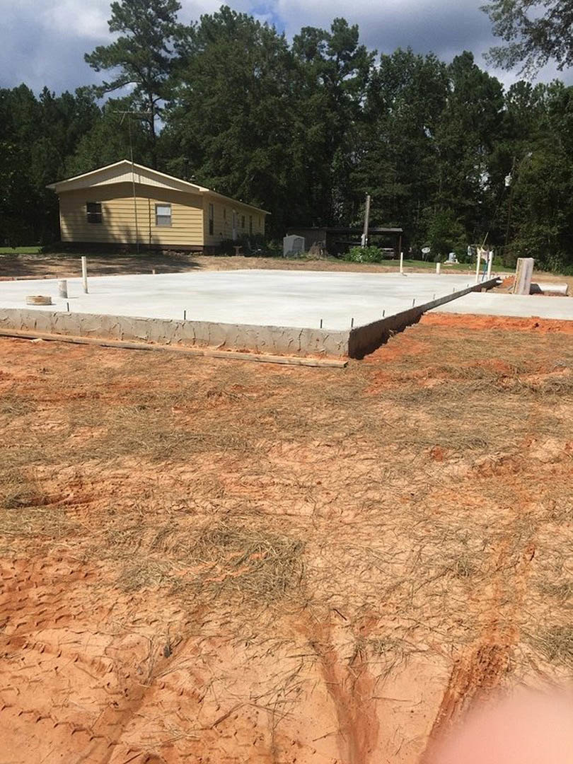 Wood-framed house under construction with exposed concrete slab, dirt and grass foreground, yellow finished home and mature trees in background