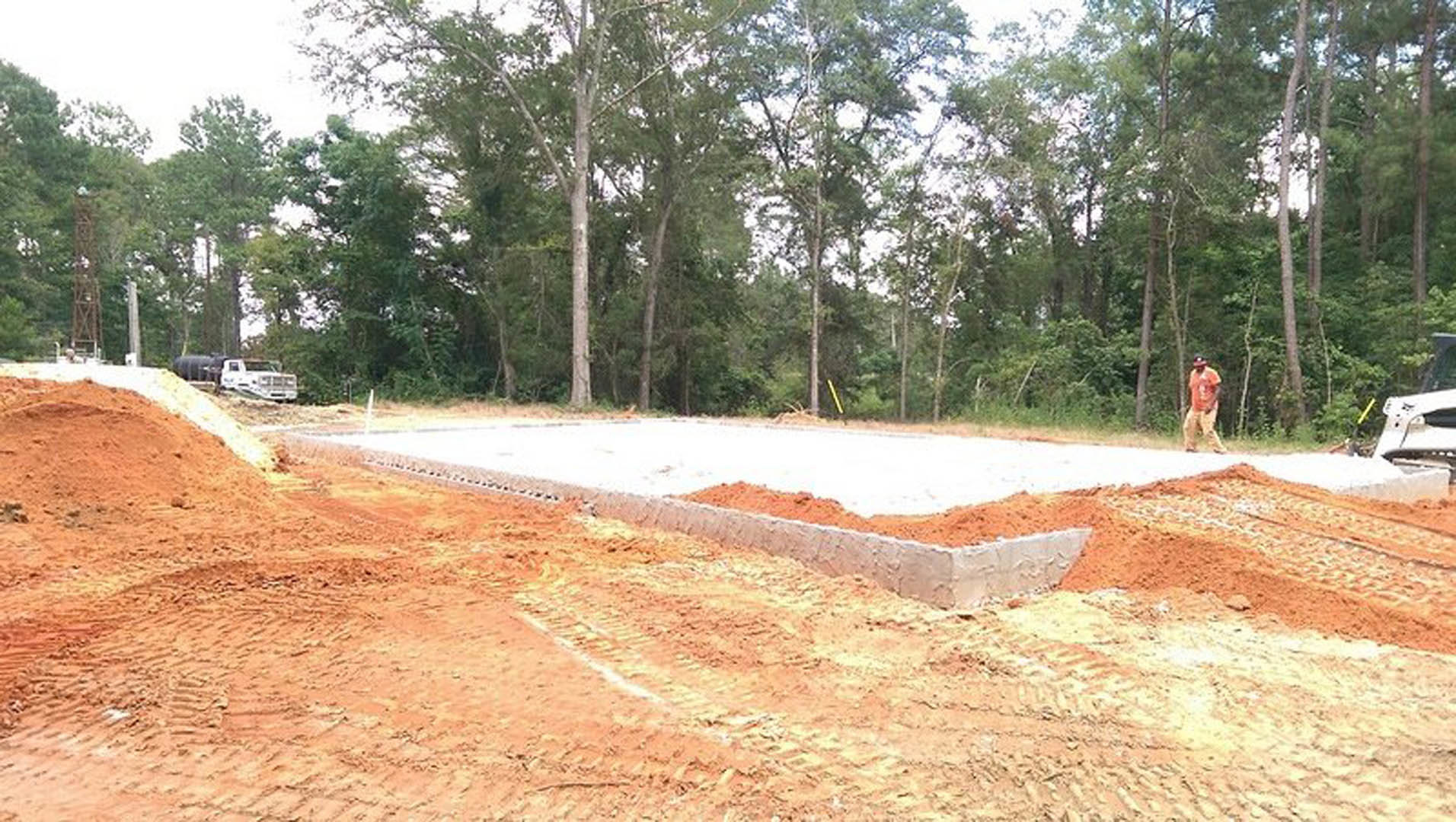 Concrete foundation under construction surrounded by dirt, trees, and construction vehicles on a clear day
