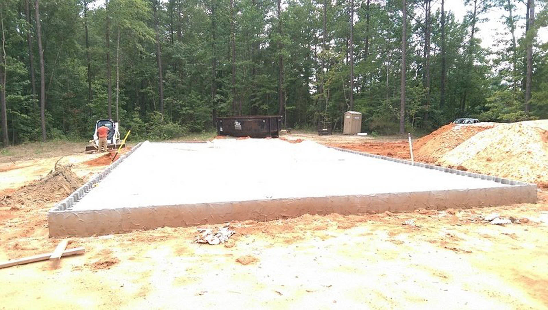 Concrete house foundation with wooden framing, surrounded by dirt and construction debris, trees in background, dumpster near wooded area.