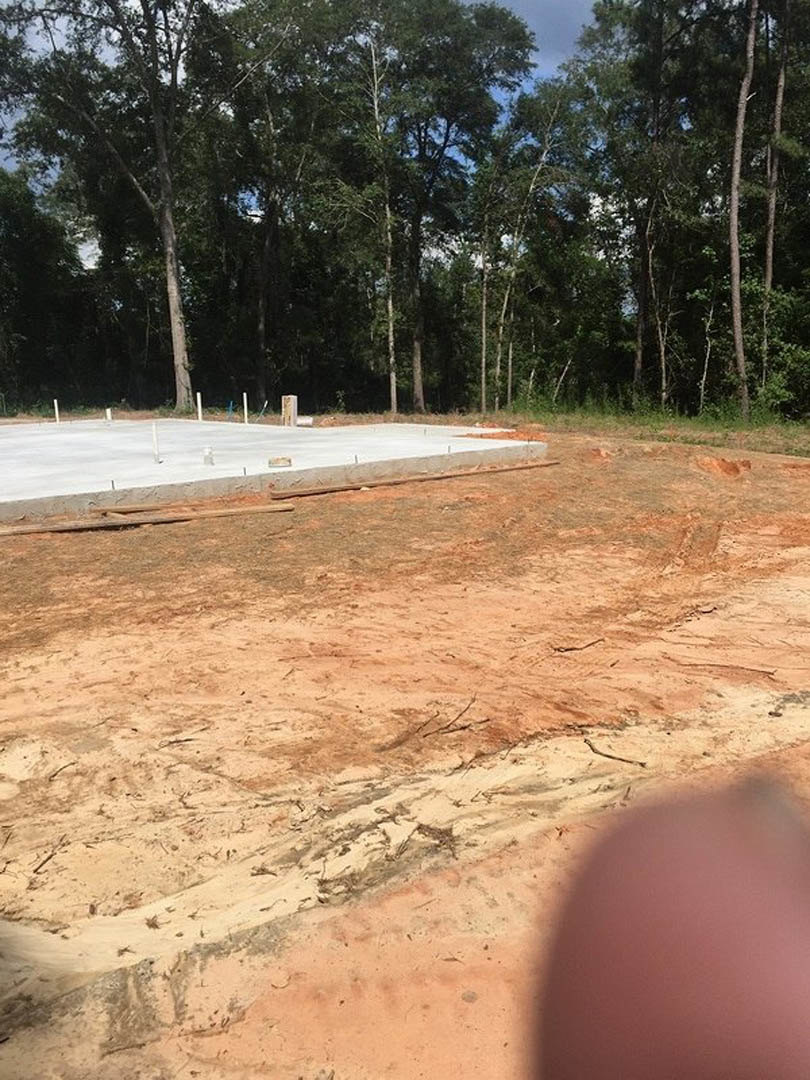 Concrete slab foundation surrounded by sandy soil and dirt, construction materials scattered on site, trees lining the background, partial view of a person at edge of frame.