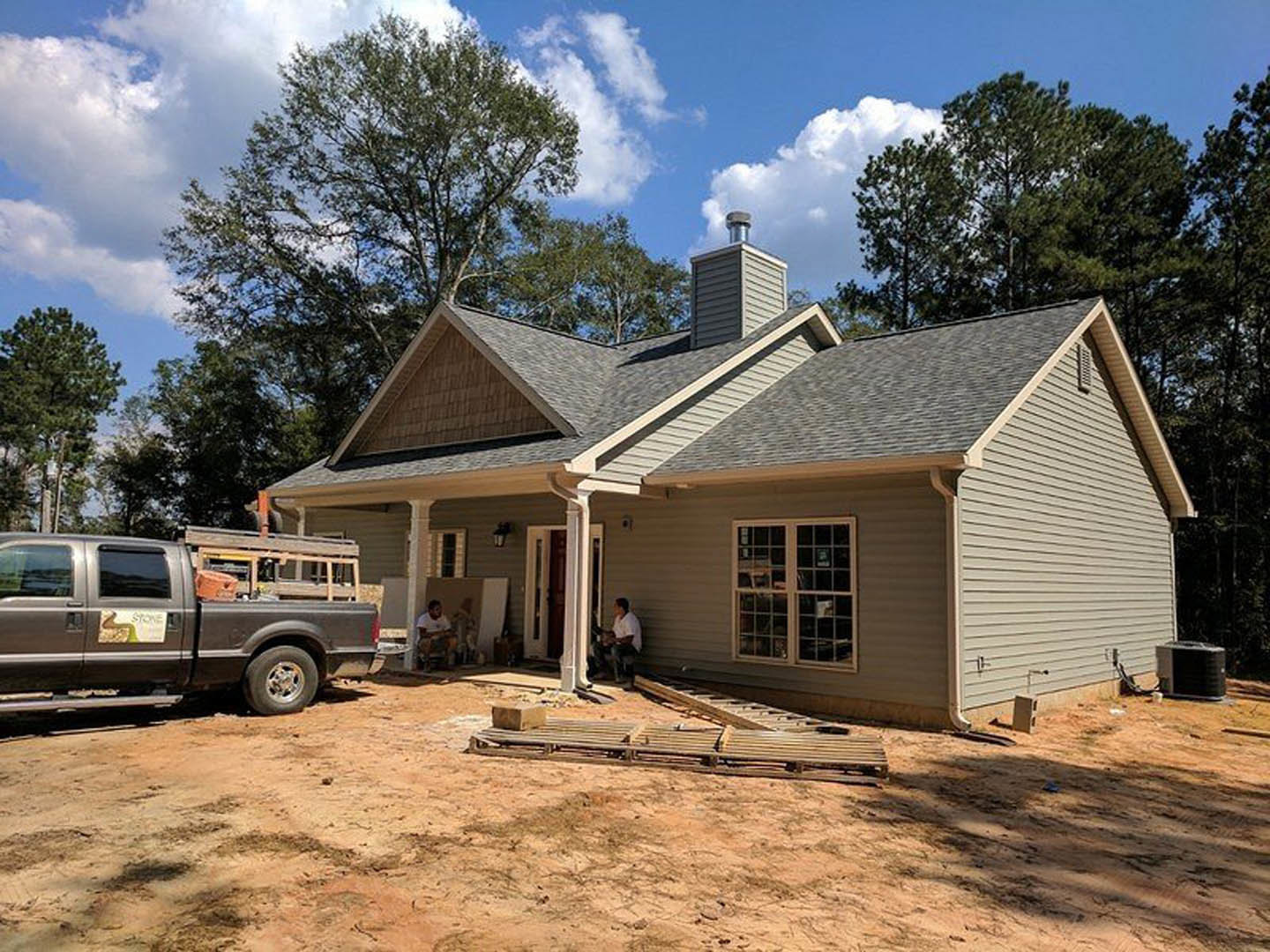 White pickup truck parked beside modern house with gray siding, large windows, and covered porch; wooden pallet and construction sign visible near entry; man seated on porch steps