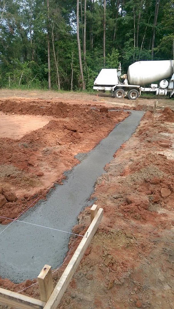 Concrete mixer on dirt near poured foundation, surrounded by trees and soil, trailer wheels visible
