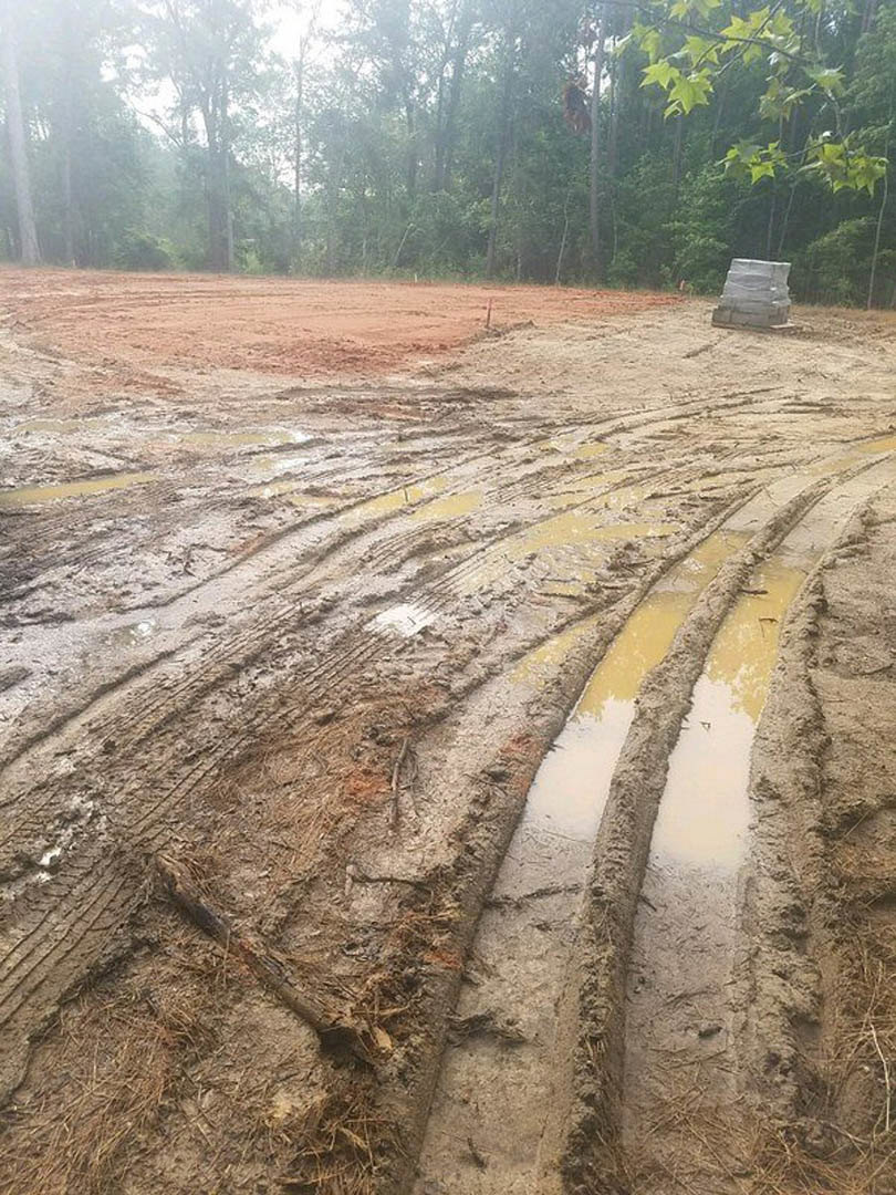 Muddy ground with deep tire tracks, standing water, scattered bricks, and a tree branch with green leaves near a foggy wooded area