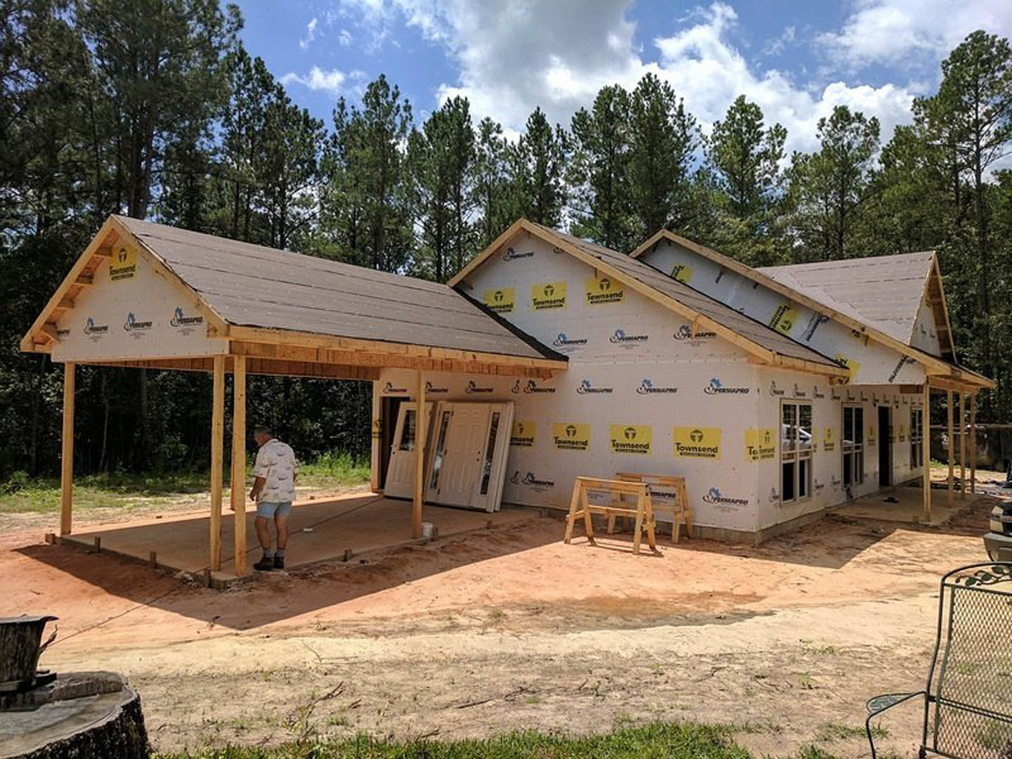 Man in white shirt and blue shorts standing on dirt in front of partially constructed house with exposed framing, white door with glass windows, wooden sawhorse, and metal chair