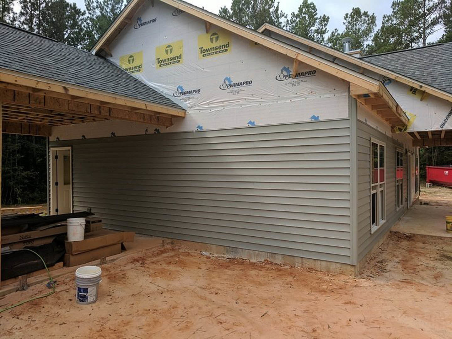 Framed house under construction with attached garage, exposed lumber, white siding, construction materials including buckets and containers on ground, unfinished door, logo sign on
