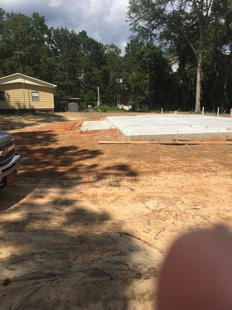 Framed house under construction with exposed roof, pickup truck parked on dirt driveway, concrete foundation, temporary fencing, scattered building materials, trees and clouds in