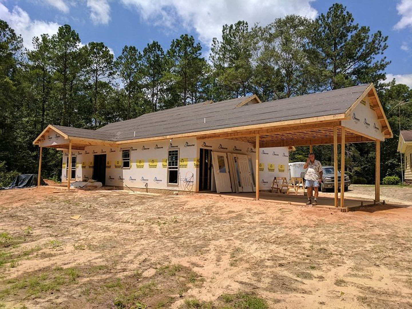 Man in shorts and white shirt standing on dirt lot in front of partially built house with exposed framing, multi-pane windows, and doors; Gordon House visible in background among