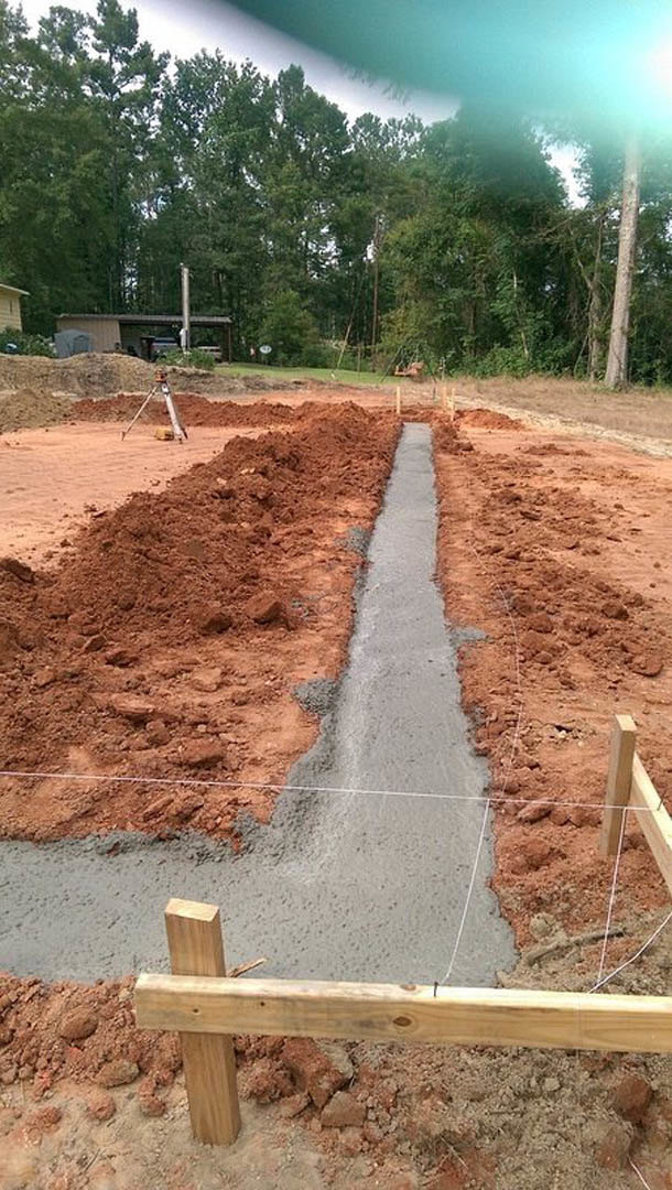 Long concrete trench running through a dirt yard, bordered by wooden boards and wire, with scattered trees and cloudy sky in the background.