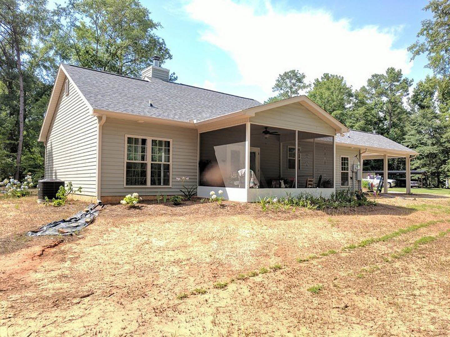 White two-story house with covered front porch, black square column, large windows, small tree and patch of dirt in landscaped yard, blue sky overhead.