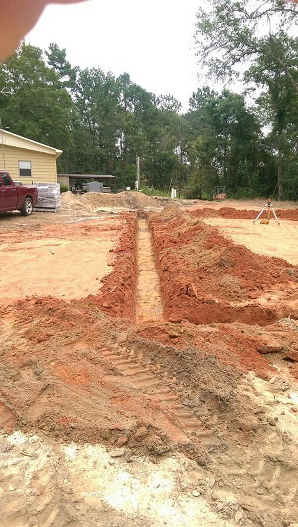 Yellow house with white trim, red car parked in driveway, dirt trench in foreground, mature trees and blue sky in background