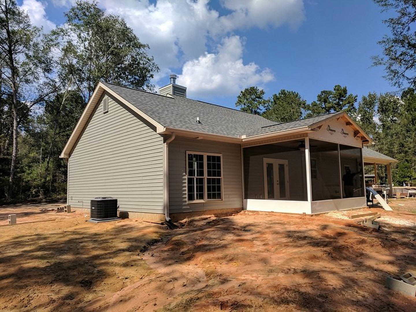 Wood-framed house under construction with covered porch, exposed beams, unfinished siding, large window, dirt yard, cloudy sky overhead