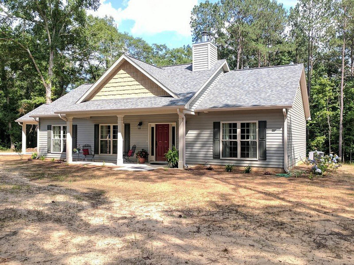 Red front door with white trim, small porch with a chair and potted plant, white cottage visible in background, green yard and mature trees surrounding home.