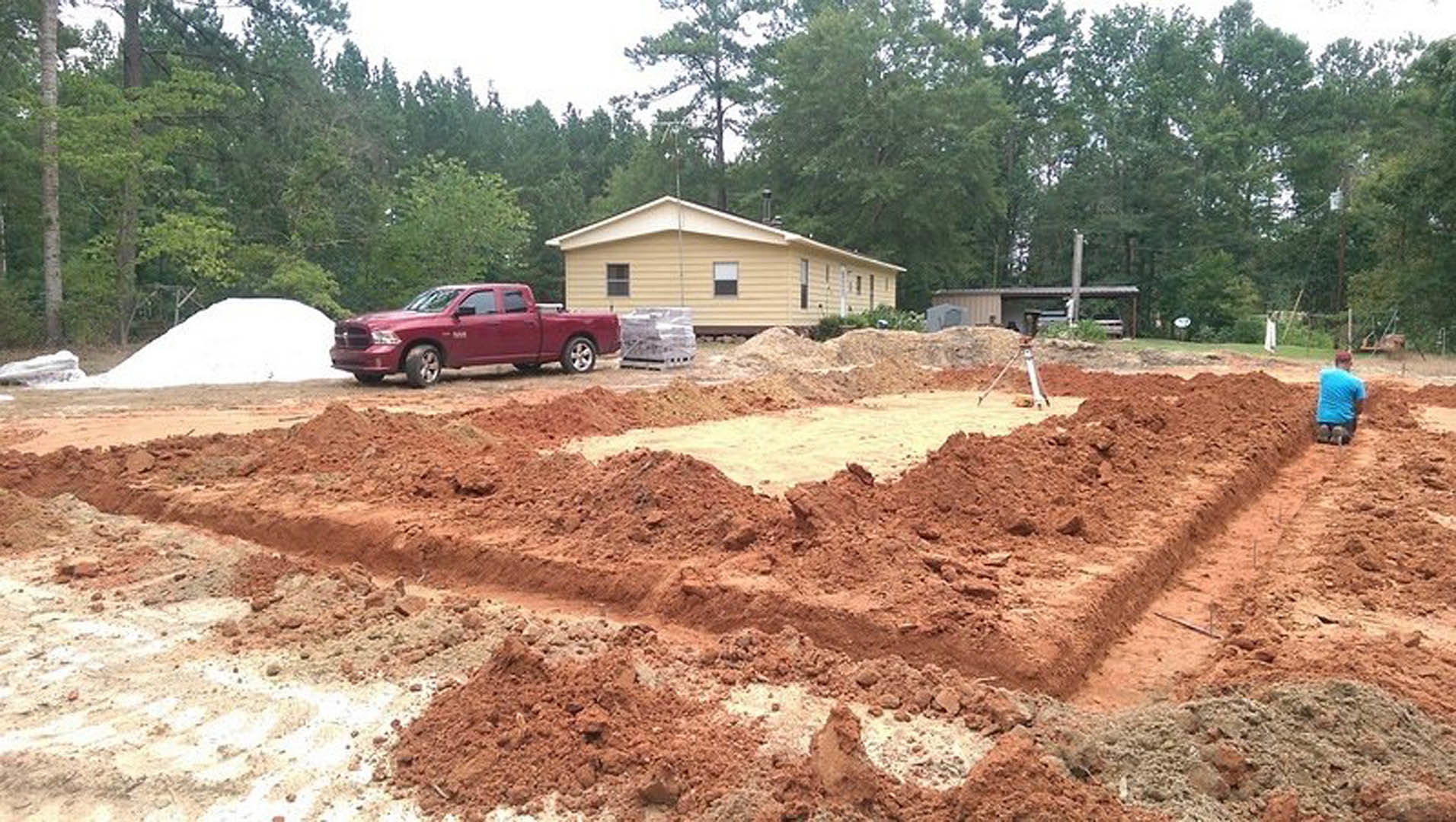 Red pickup truck parked on dirt driveway beside yellow house with white-trimmed window, surrounded by trees under blue sky.