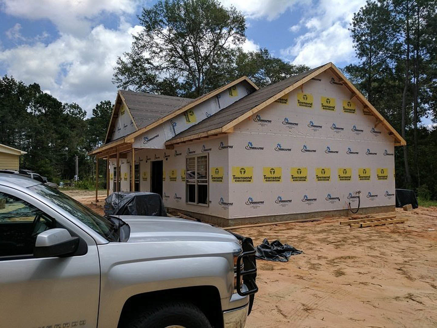 Framed house under construction with exposed plywood walls and roof, pickup truck parked on dirt driveway, cloudy sky overhead