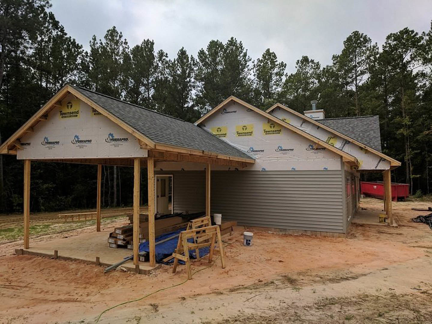 Framed house under construction with exposed wood beams, covered porch, dirt yard, wooden chair, and red container near train tracks