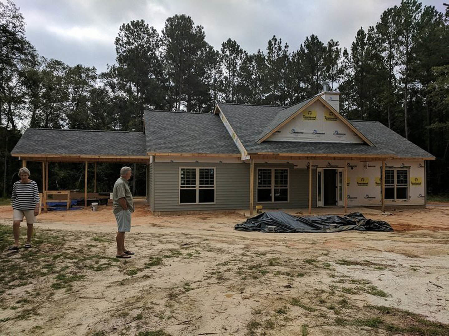 Man in striped shirt standing on dirt ground with black tarp in front of partially built house, exposed framing and unfinished windows visible, surrounded by trees under cloudy sky