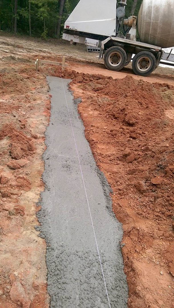 Freshly poured concrete road bordered by dirt and grass, large cement mixer truck parked nearby, close-up details of truck tires and construction materials visible.