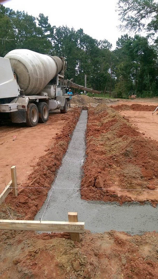 Concrete mixer truck parked on dirt construction site, surrounded by soil, wooden boards, and temporary fencing, with trees and sky in background