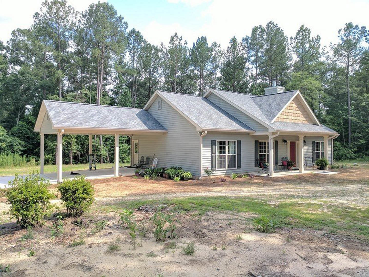 Covered porch with white roof, white-framed windows, bush in foreground, dirt patch, trees and small white cottage in background