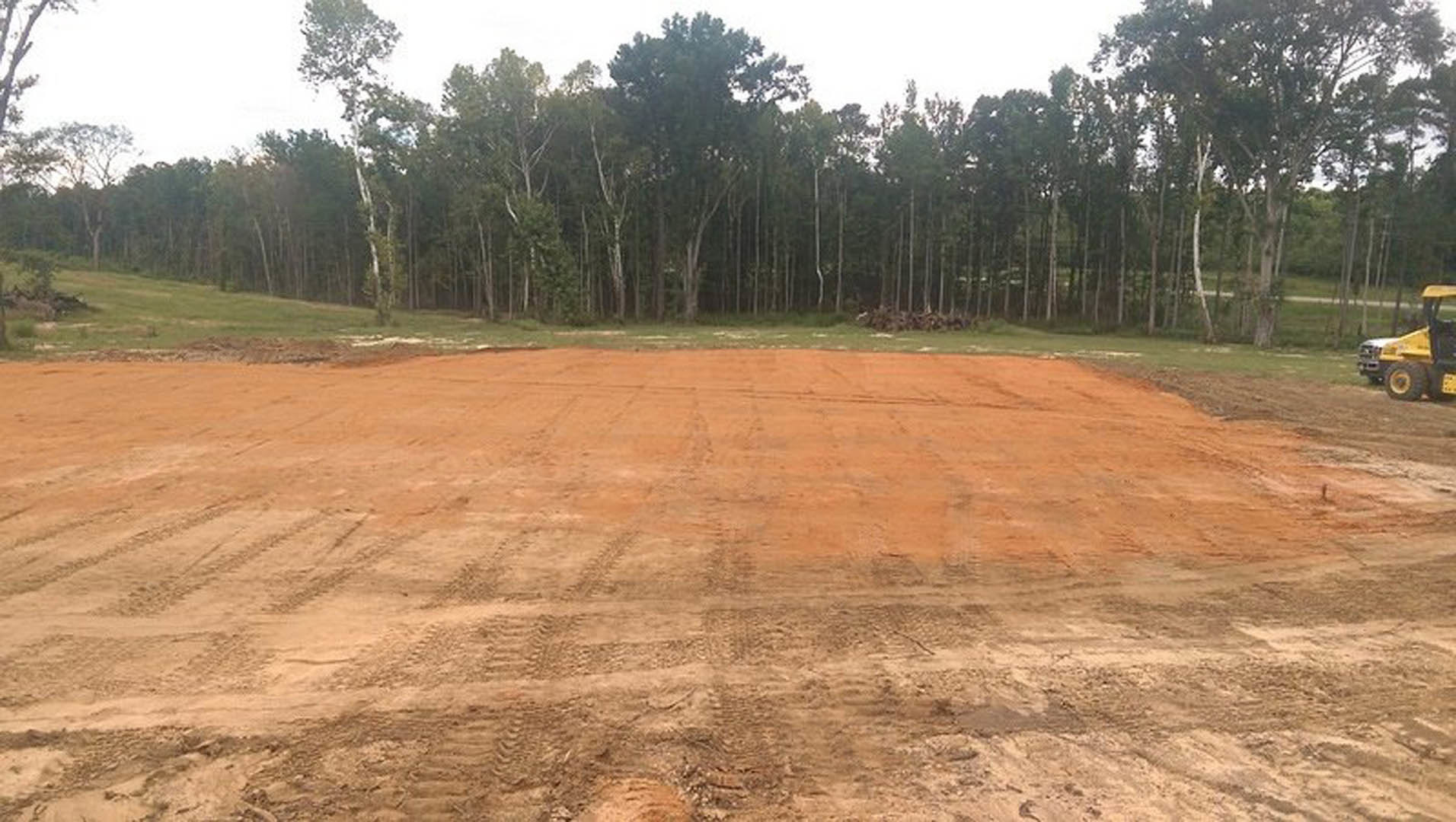Dirt field with tire tracks, yellow truck parked near grassy edge, group of trees in background under clear sky