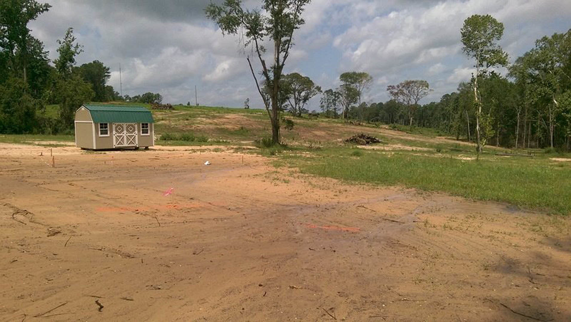 Small house with green roof and white siding set in grassy field, surrounded by tall trees with dense green leaves, under partly cloudy sky.