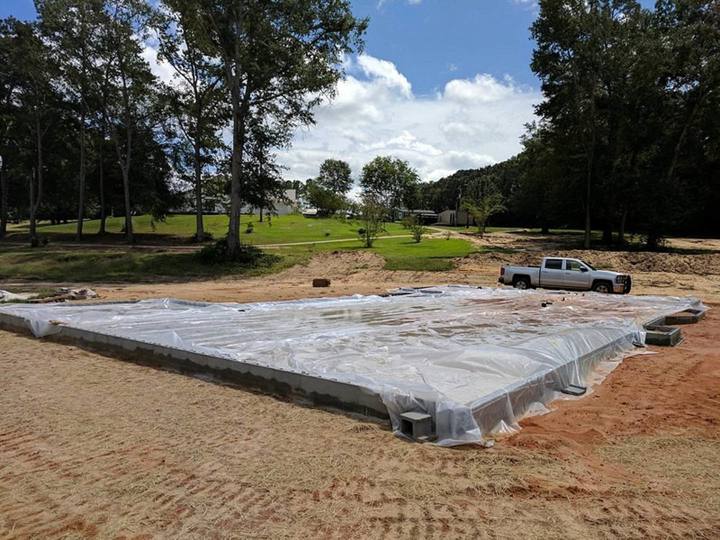 Concrete foundation covered with plastic tarp beside a white truck parked on a dirt road, surrounded by grassy field, trees, and blue sky with clouds