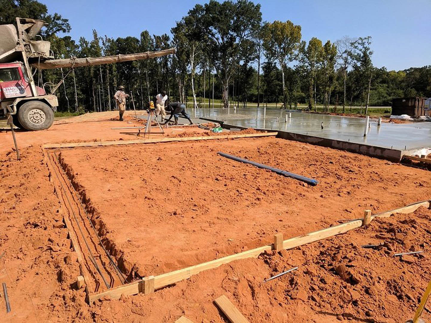 Framed custom home under construction with workers on muddy ground, pickup truck parked nearby, scattered wood planks, tire tracks in soil, trees and blue sky in background