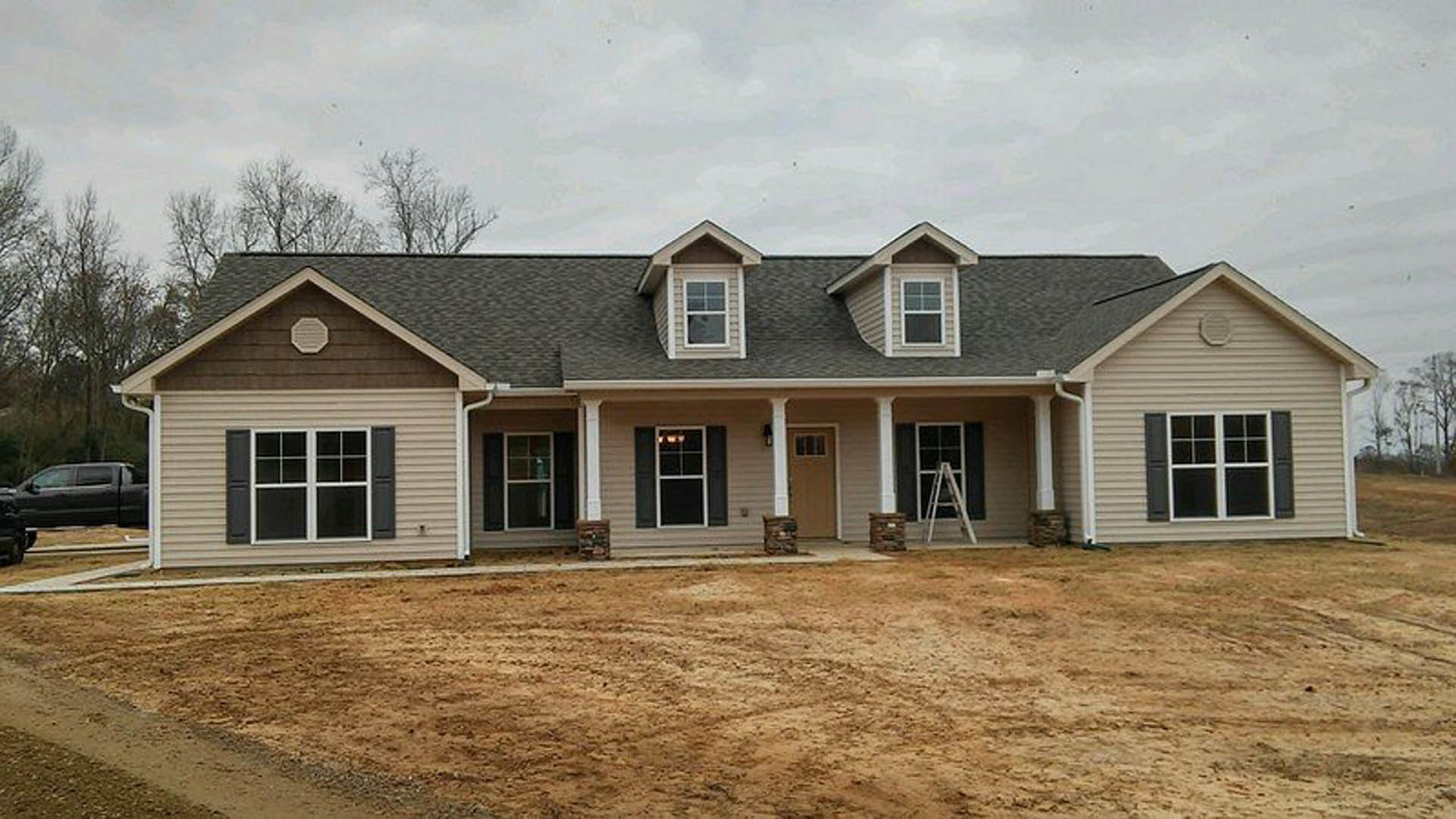 Two-story house with light siding, several rectangular windows, and a dirt yard; black pickup truck parked nearby, person walking across open ground, cloudy sky overhead.