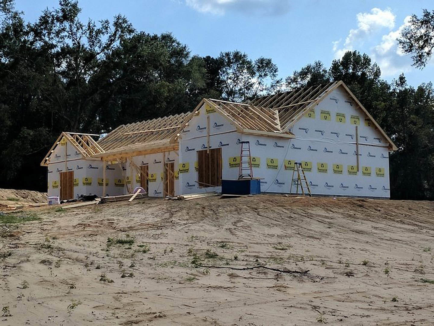 Partially built house with exposed brown wood paneling, construction materials scattered on dirt ground, ladder leaning against unfinished doorway, trees and cloudy sky in