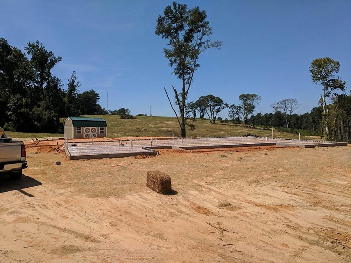 Wood-framed house under construction on a dirt lot, blue metal roof partially installed, surrounded by open field and scattered trees, truck parked nearby, hay bale visible in